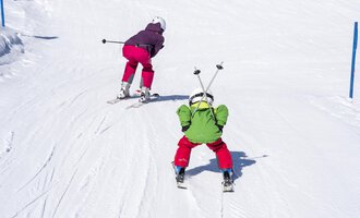 Two children are skiing on a snowy slope. One child is wearing a green suit and the other a purple suit. | © Kleinwalsertal Tourismus | Dominik Berchtold