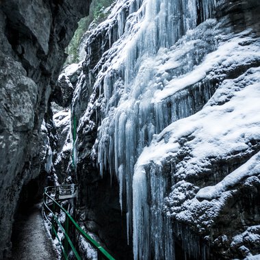 Eisiges Gestein in der Breitachklamm im Winter mit gefrorenen Wasserfällen und Schneeschicht, ein schmaler Weg mit Geländer führt durch die Klamm. | © Kleinwalsertal Tourismus eGen • Fotograf: Dominik Berchtold