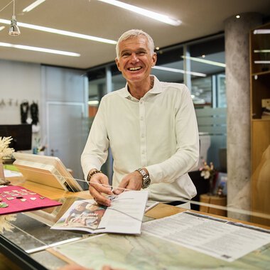 Mann im Büro hinter dem Kundenservice-Schalter, lächelnd, mit Unterlagen und Kasse. | © Kleinwalsertal Tourismus eGen • Fotograf: Oliver Farys