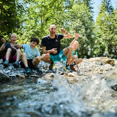 A lively group of four people, including children and an adult, sitting by a creek in a green forest, enjoying playing in the water. | © Kleinwalsertal Tourismus eGen • Photographer: Oliver Farys