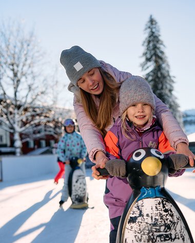 Kinder beim Eislaufen auf einem winterlichen Eisplatz in Riezlern im Kleinwalsertal, Sonnenlicht und schneebedeckte Bäume im Hintergrund. | © Kleinwalsertal Tourismus eGen • Fotograf: Oliver Farys
