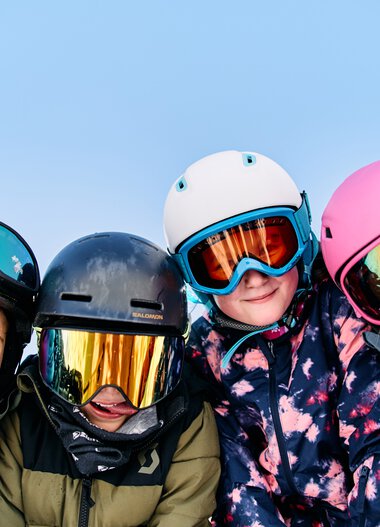 Four children in winter clothing and helmets, smiling and posing in the snow while skiing. | © Kleinwalsertal Tourismus eGen • Photographer: Oliver Farys