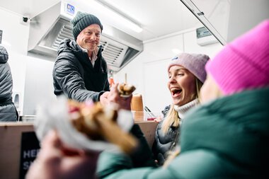 Friendly snowman gathering in Kleinwalsertal where a man serves food to guests who are laughing and having fun. | © Kleinwalsertal Tourismus eGen • Photographer: Oliver Farys