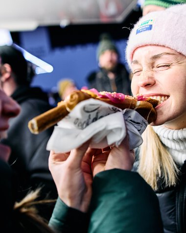 Two women at the snowman gathering in Kleinwalsertal, one is eating a piece of pizza, both are laughing and wearing winter clothing. | © Kleinwalsertal Tourismus eGen • Photographer: Oliver Farys